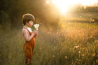 little boy in a field in orange overalls blowing dandelions