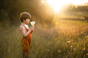 little boy in a field in orange overalls blowing dandelions