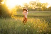 little boy in a field in orange overalls with a hat smiling