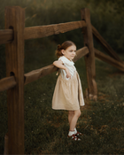 A young girl with a ponytail stands outdoors leaning against a rustic wooden fence in a grassy field. She is wearing a white collared shirt under a light beige linen pinafore-style dress and brown sandals.