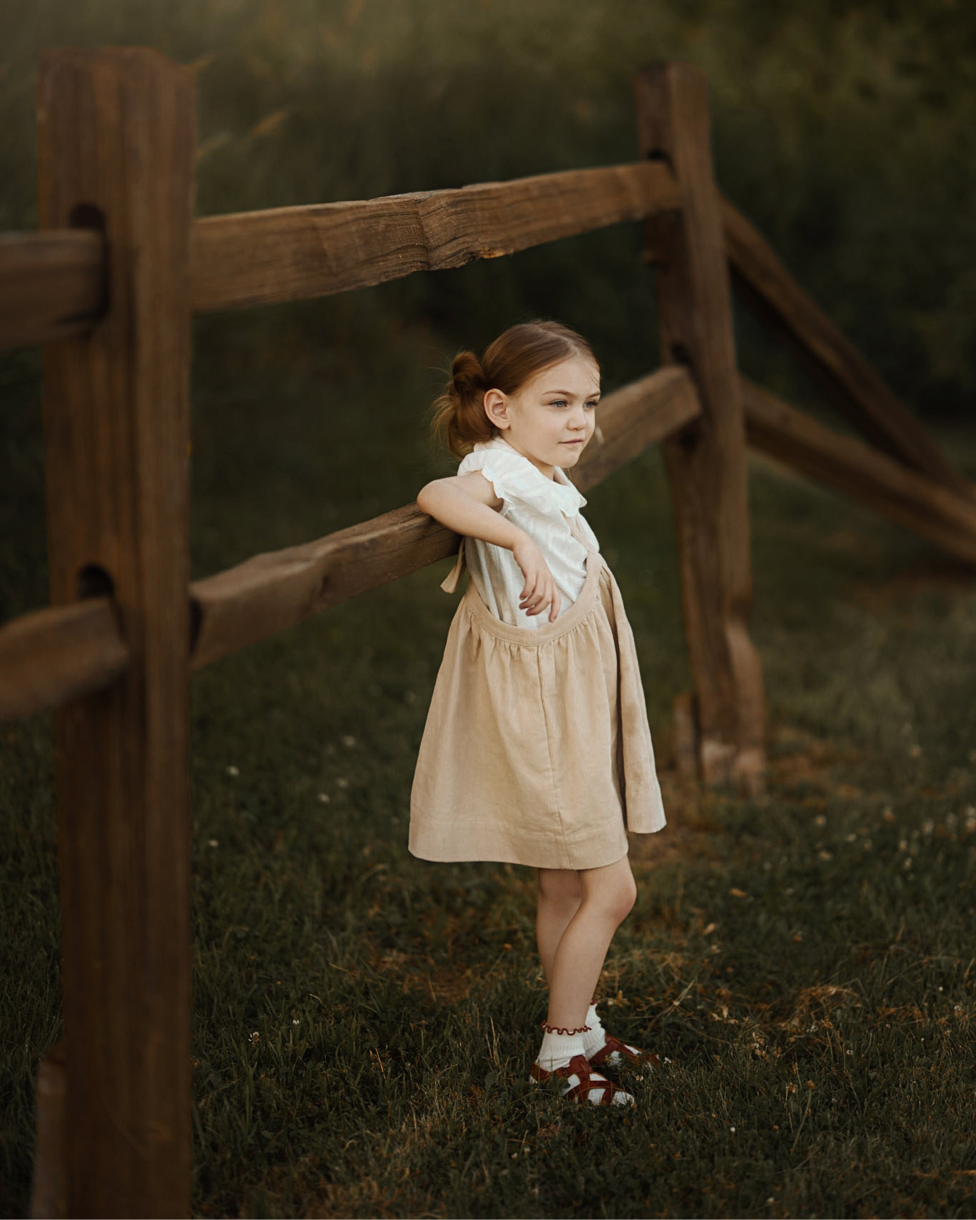 A young girl with a ponytail stands outdoors leaning against a rustic wooden fence in a grassy field. She is wearing a white collared shirt under a light beige linen pinafore-style dress and brown sandals.