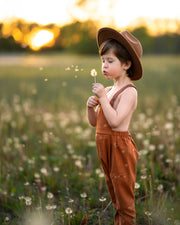 little boy in a field in orange overalls blowing dandelions 