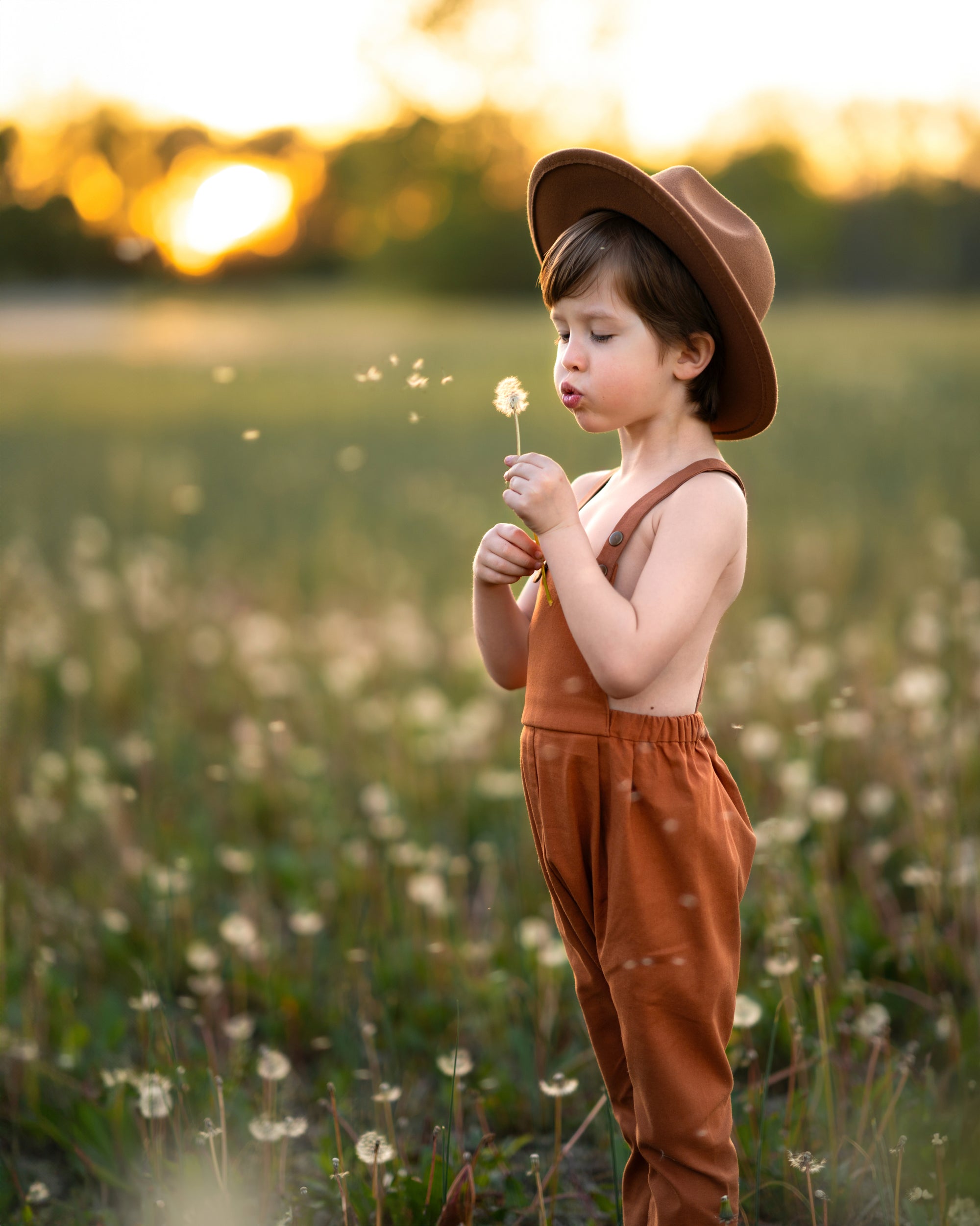 little boy in a field in orange overalls blowing dandelions 