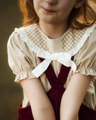 A close-up of a child wearing a cream and tan gingham top with a large white ruffled collar and bow, layered under a dark red corduroy pinafore dress.