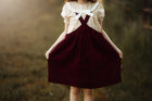 A young girl wearing a cream short-sleeve blouse with a large white bow at the neckline and dark burgundy corduroy pinafore overalls, holding the skirt out in an outdoor field.