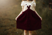 A young girl wearing a cream short-sleeve blouse with a large white bow at the neckline and dark burgundy corduroy pinafore overalls, holding the skirt out in an outdoor field.