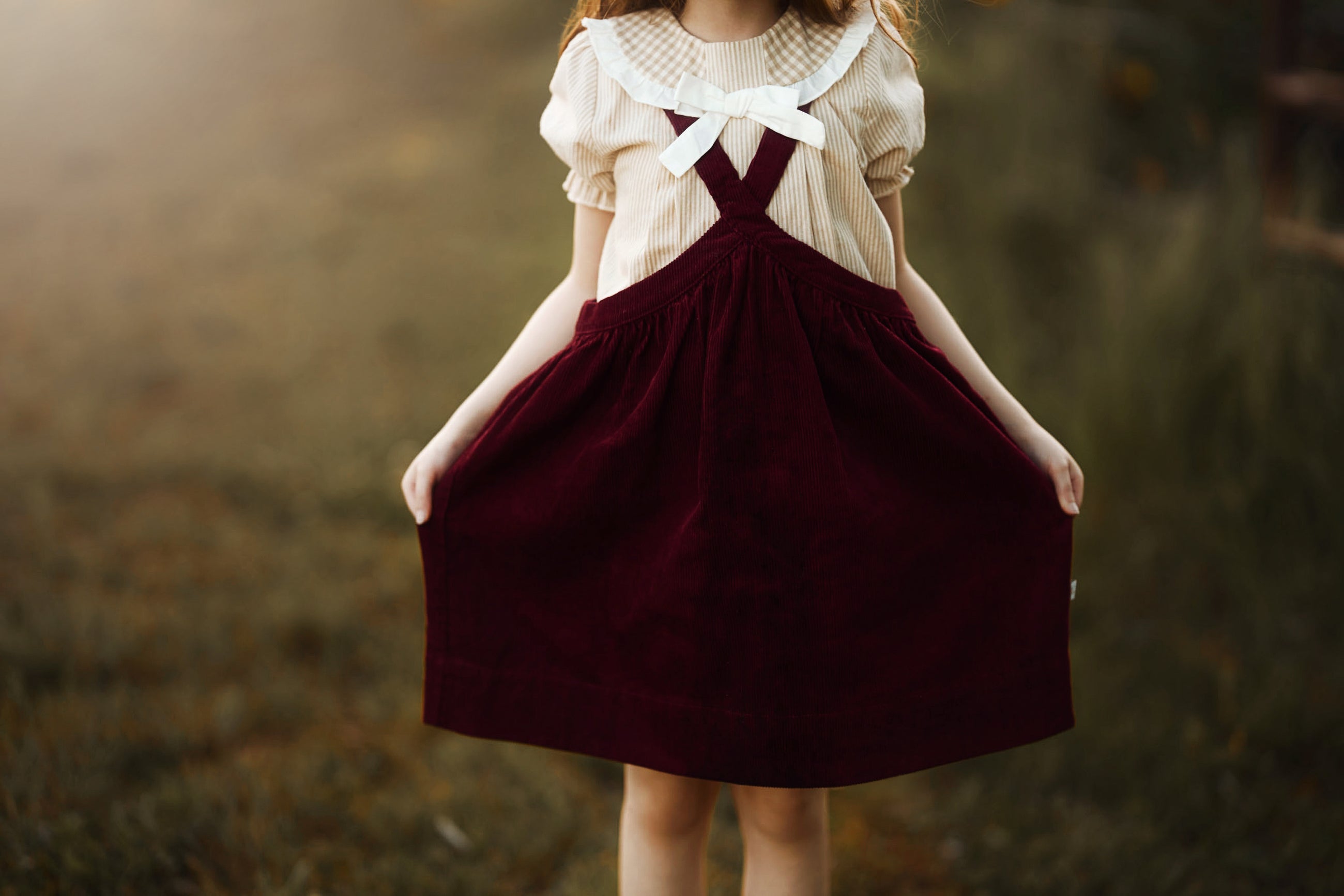 A young girl wearing a cream short-sleeve blouse with a large white bow at the neckline and dark burgundy corduroy pinafore overalls, holding the skirt out in an outdoor field.