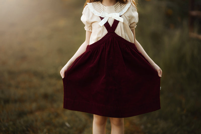 A young girl wearing a cream short-sleeve blouse with a large white bow at the neckline and dark burgundy corduroy pinafore overalls, holding the skirt out in an outdoor field.