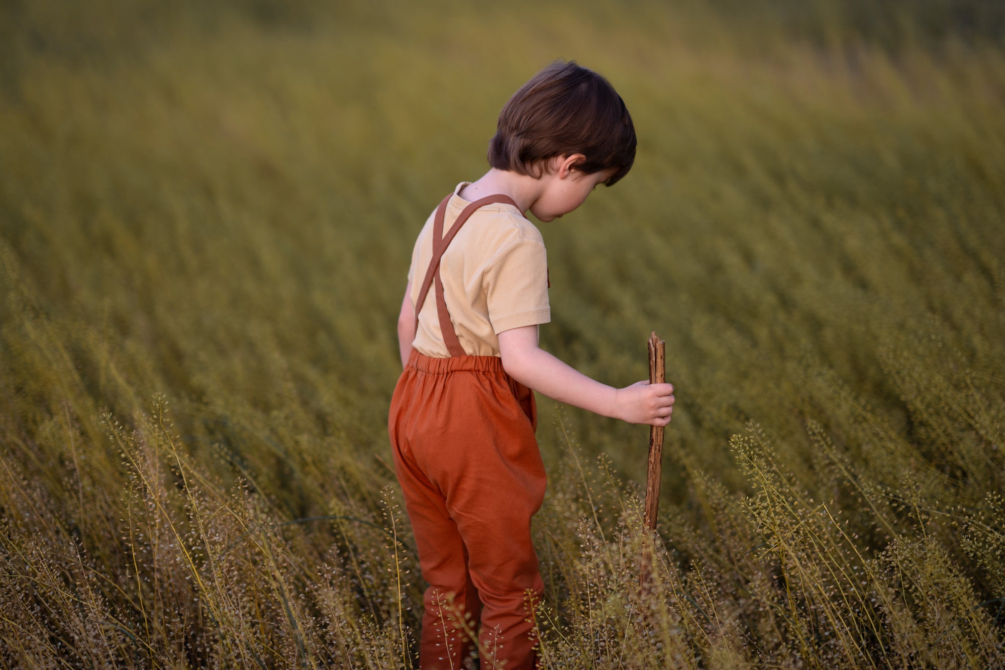 little boy in a field in orange overalls playing with a stick