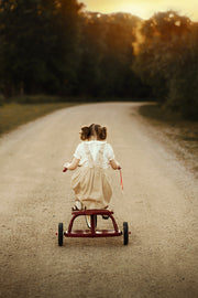 Two young children riding a small red tricycle down a dirt road, photographed from behind during golden hour with trees in the background.