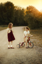 Two young girls outdoors on a dirt path at sunset. One girl stands wearing a cream top and dark red pinafore dress, while the other sits on a small red tricycle wearing a cream dress.