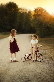 Two young girls outdoors on a dirt path at sunset. One girl stands wearing a cream top and dark red pinafore dress, while the other sits on a small red tricycle wearing a cream dress.