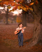 Child holding a guitar in an autumn setting with trees and fallen leaves.