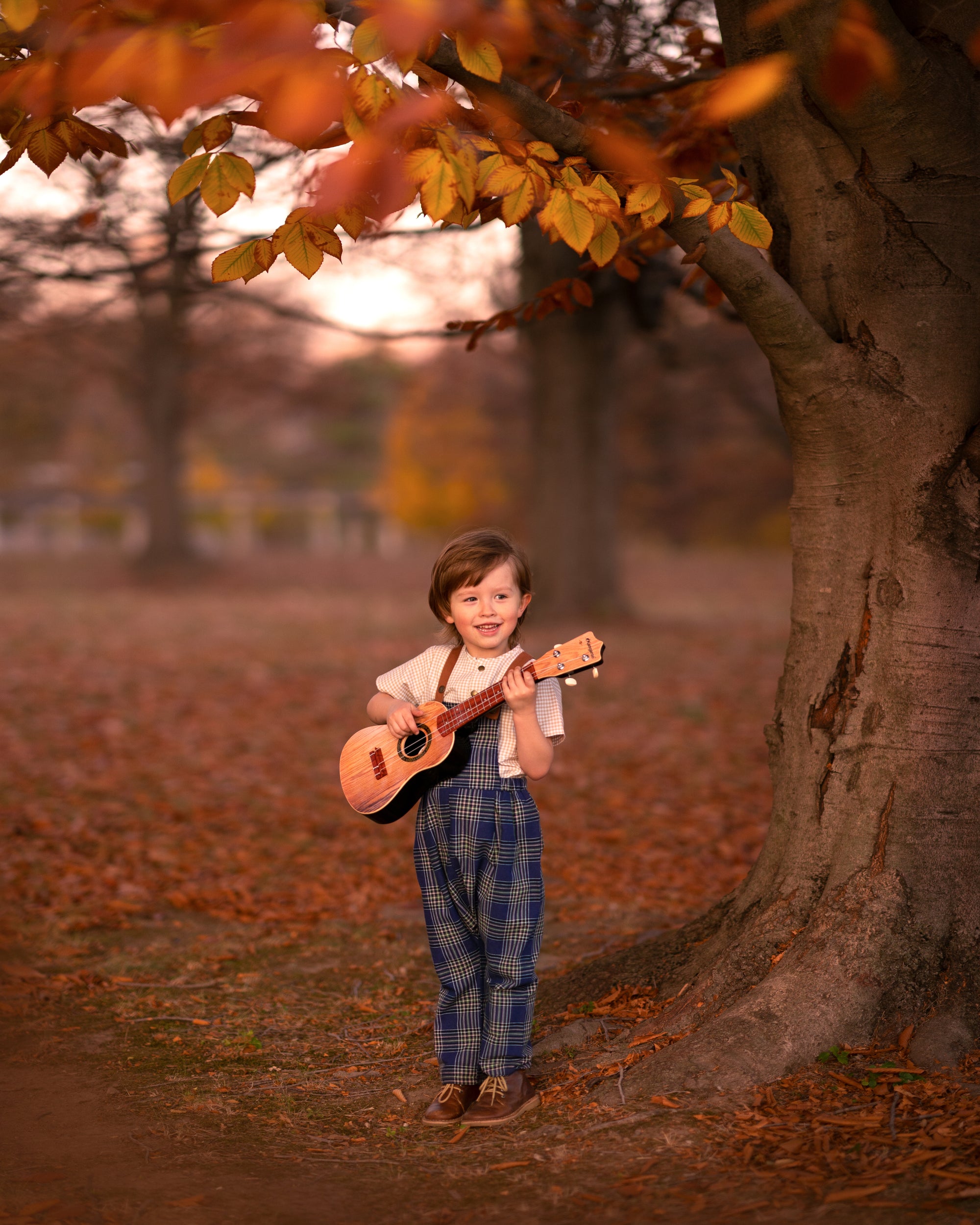 Child holding a guitar in an autumn setting with trees and fallen leaves.