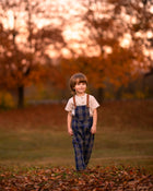 Child wearing plaid overalls standing in a field with trees in the background
