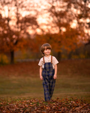 Child wearing plaid overalls standing in a field with trees in the background