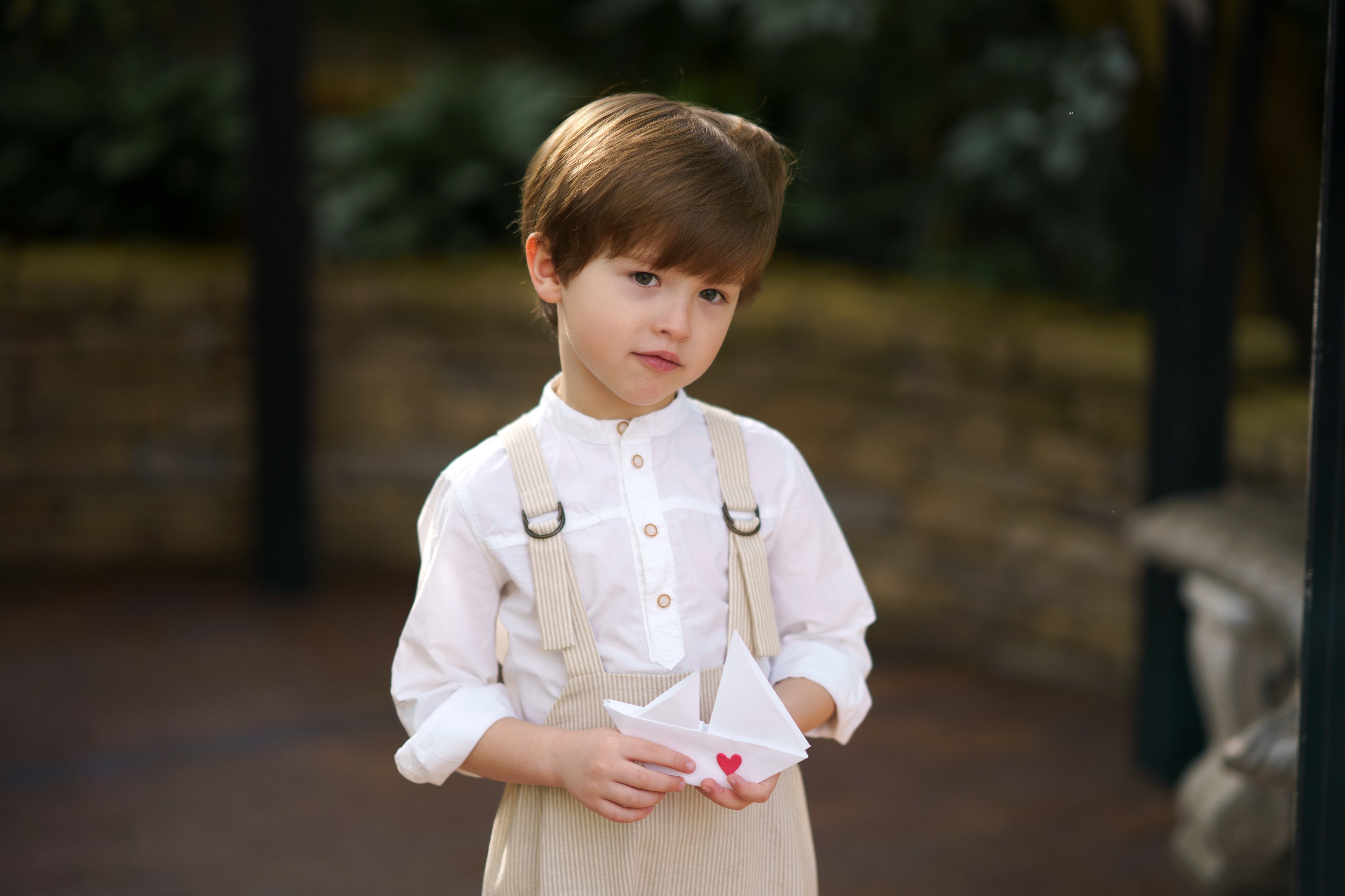 Little boy showcasing details of the Grow-With-Me Romper - Beige Stripes, highlighting the fully adjustable straps for a customizable fit, the classic timeless beige stripes pattern, and the soft, breathable fabric that ensures comfort throughout the day, representing stylish versatility in sustainable children's attire.