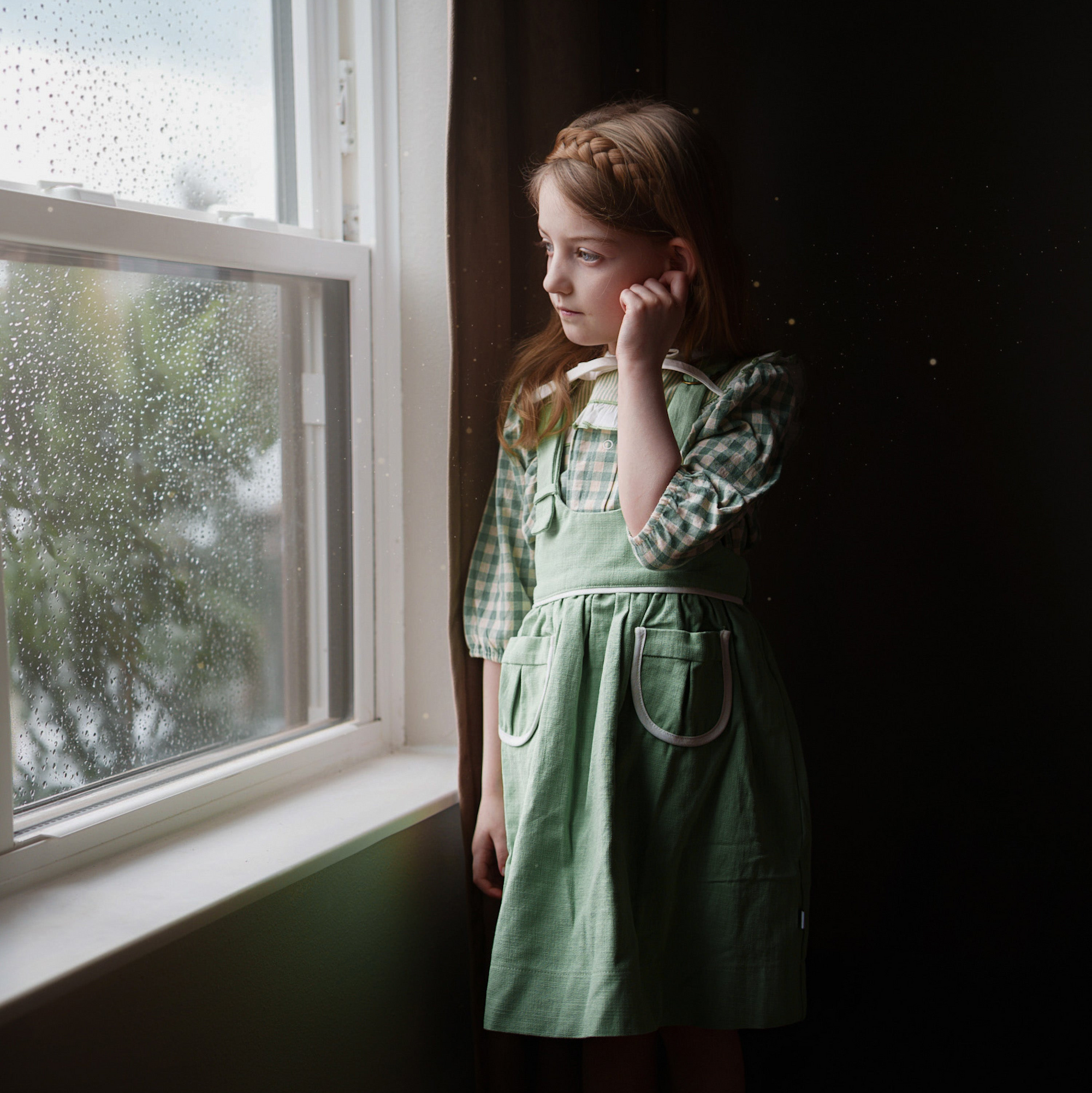 Young girl in a green dress standing by a window with rain outside