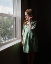 Young girl in a green dress standing by a window with rain outside