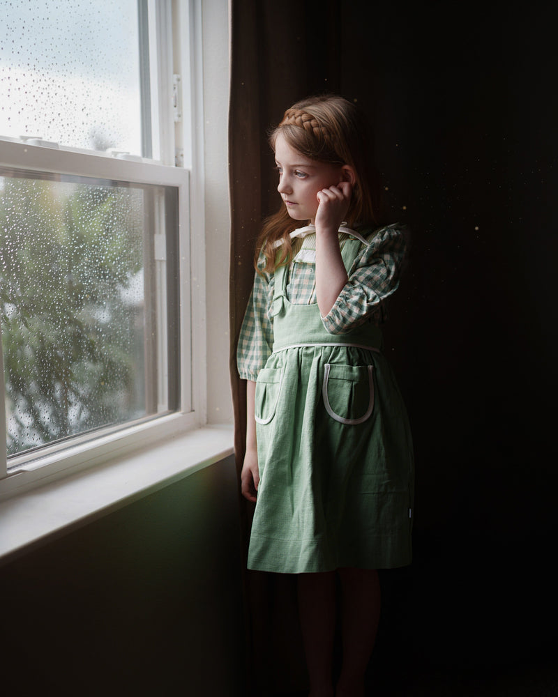 Young girl in a green dress standing by a window with rain outside