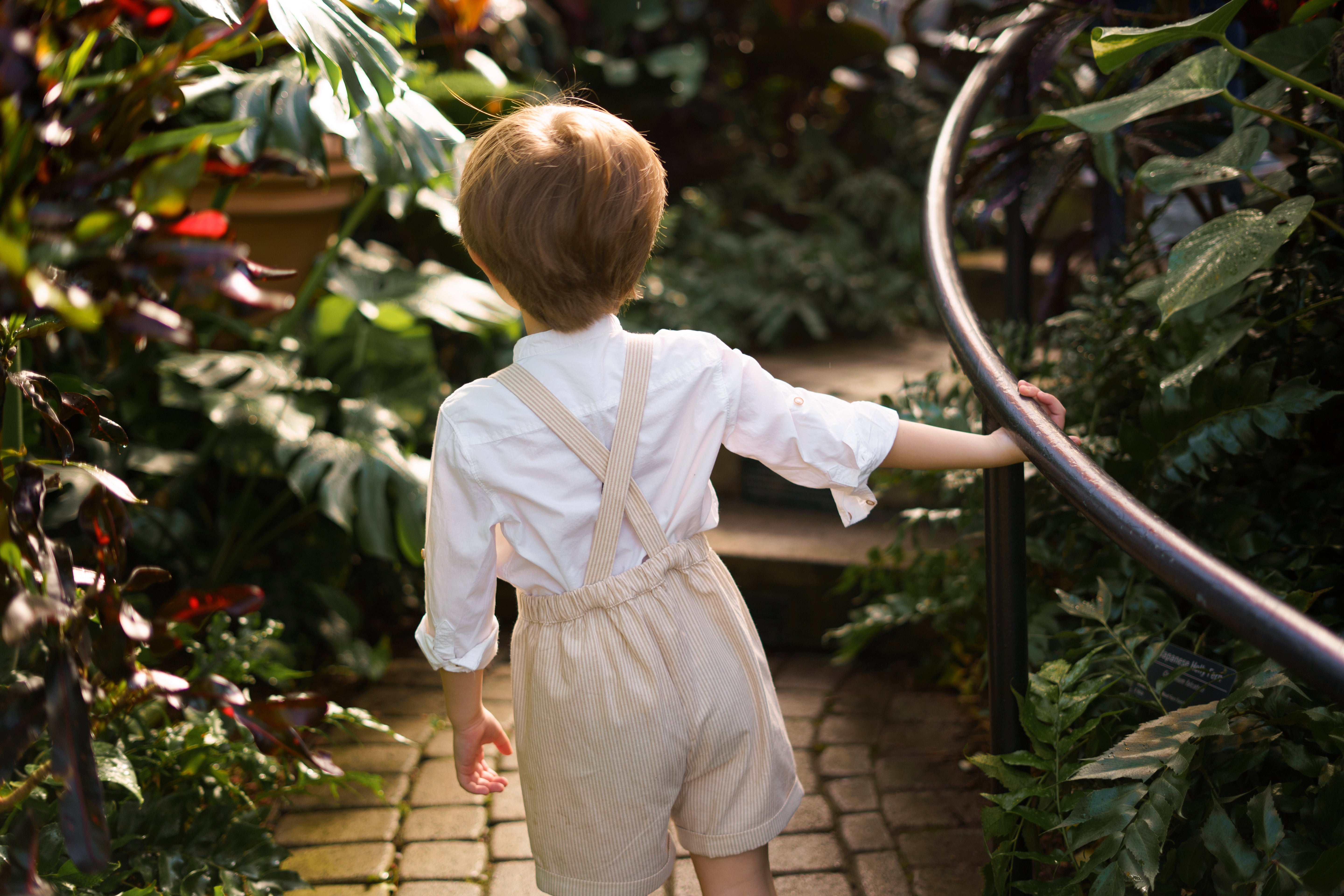 The back of a little boy wearing a beige stripes Grow-With-Me Romper, walking joyfully in a green house, highlighting the adjustable straps and comfortable fit.