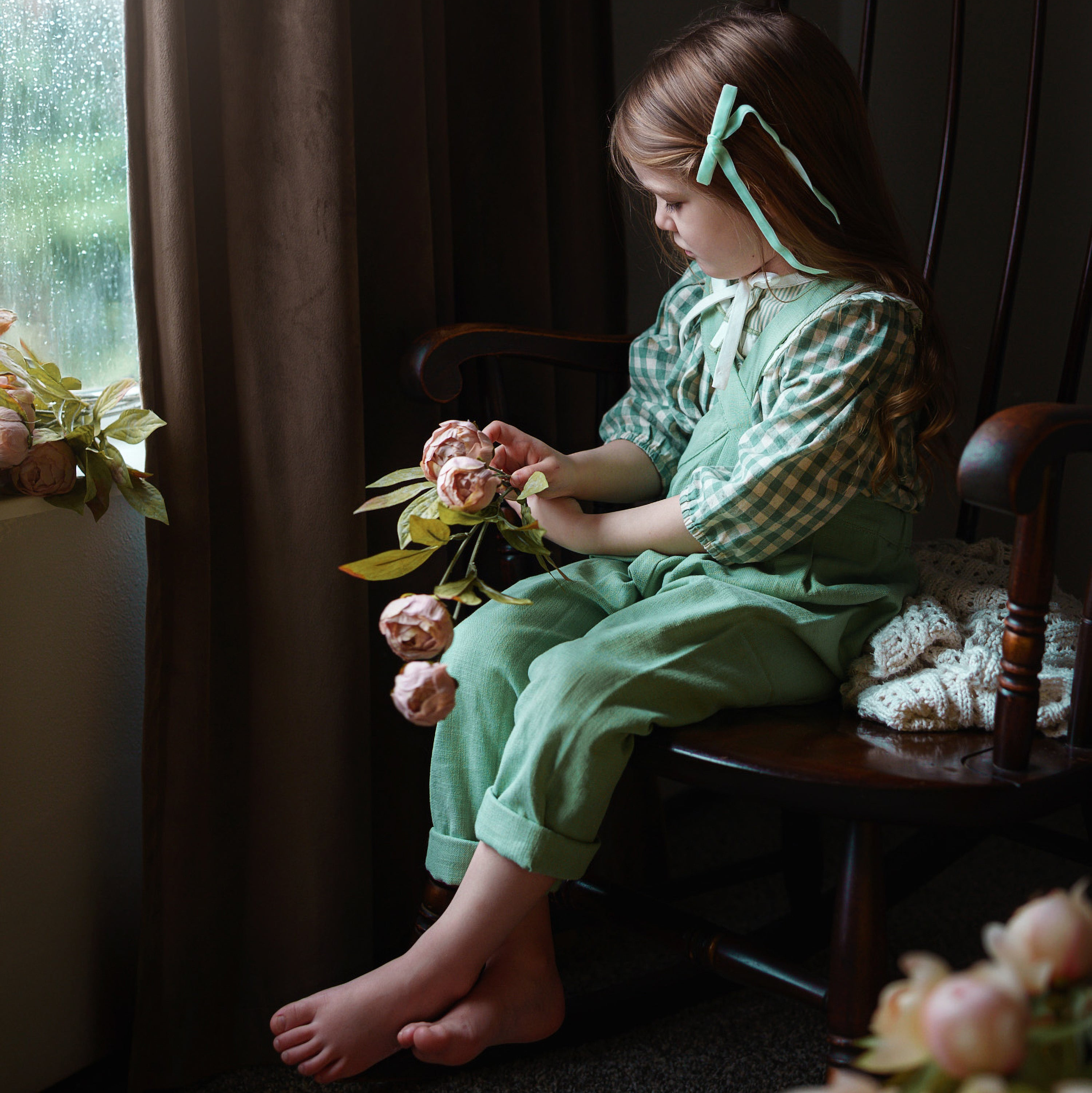 Child in a green dress sitting in a rocking chair by a window with flowers.