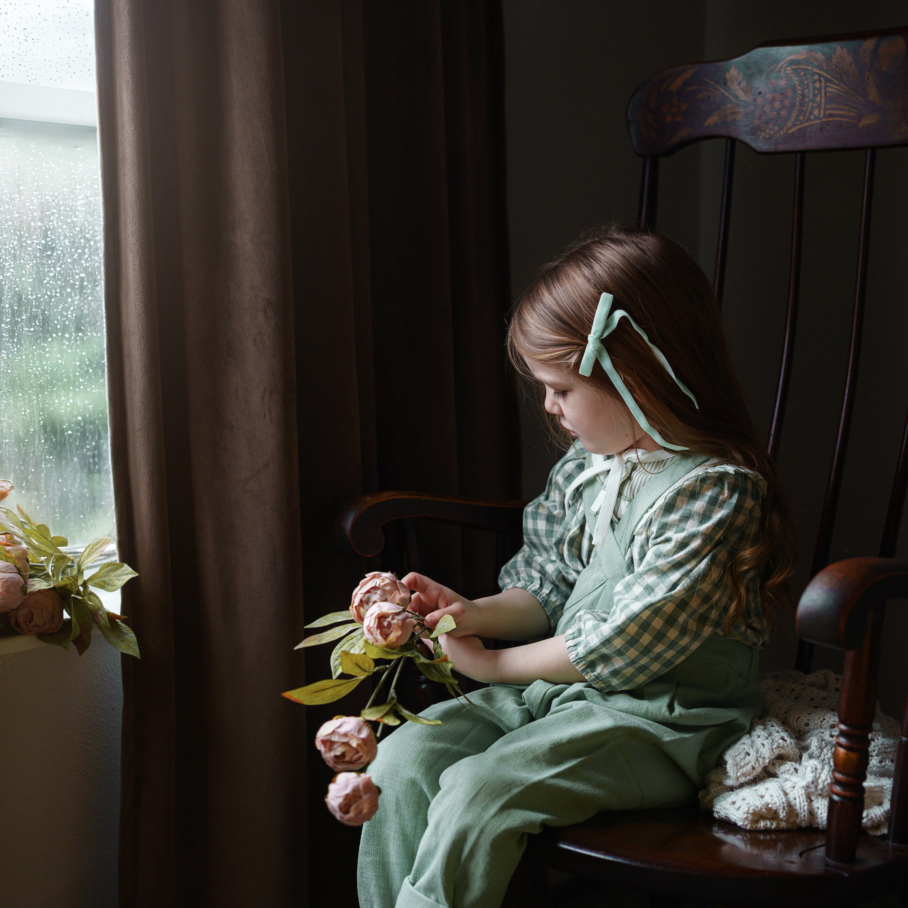Child sitting in a rocking chair holding flowers by a window with rain outside