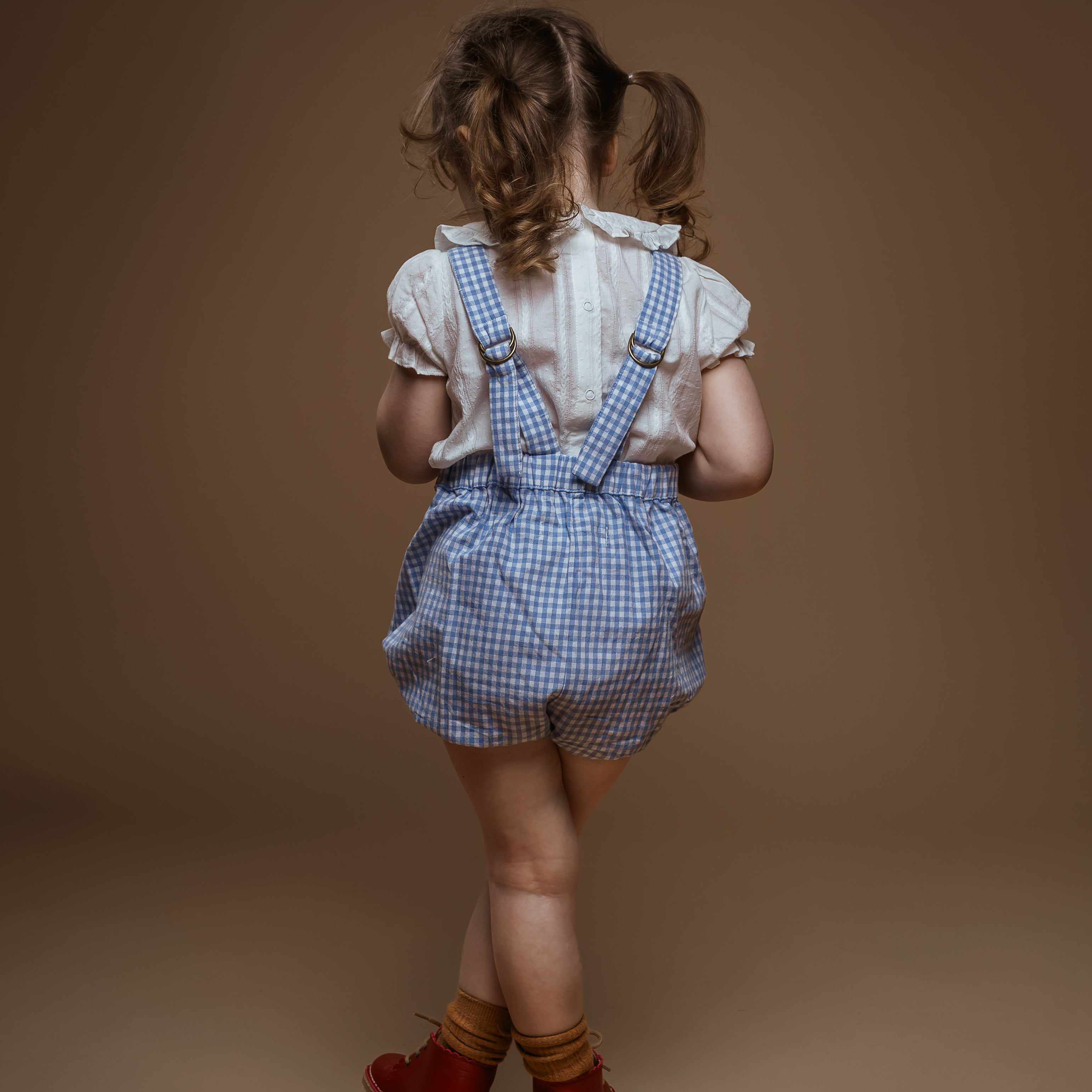 Child wearing blue checkered overalls and red shoes on a brown background