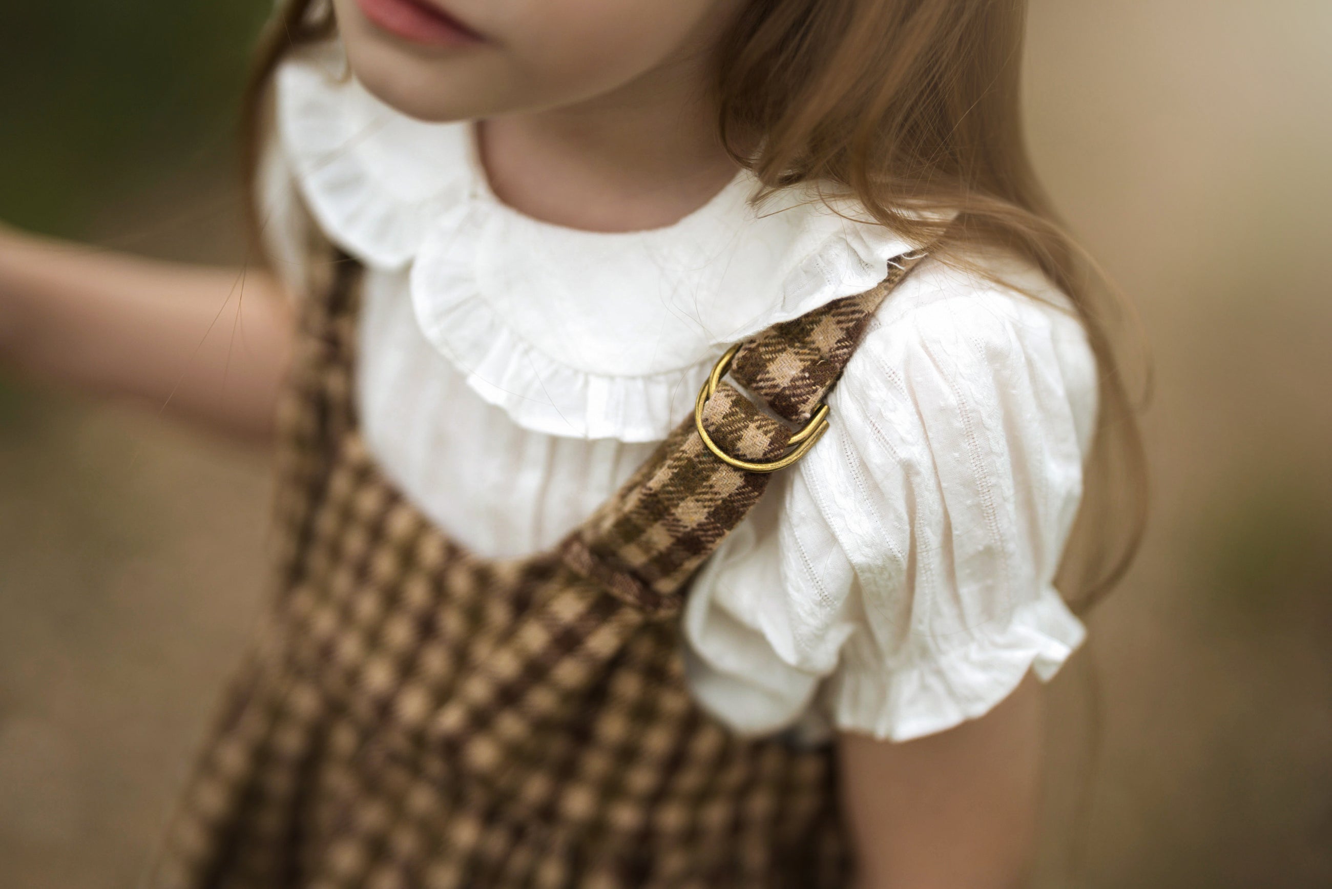 A close-up lifestyle shot of a young girl wearing a white ruffled blouse and brown checkered pinafore overalls with a gold ring detail on the strap, set against a soft, blurred outdoor background.