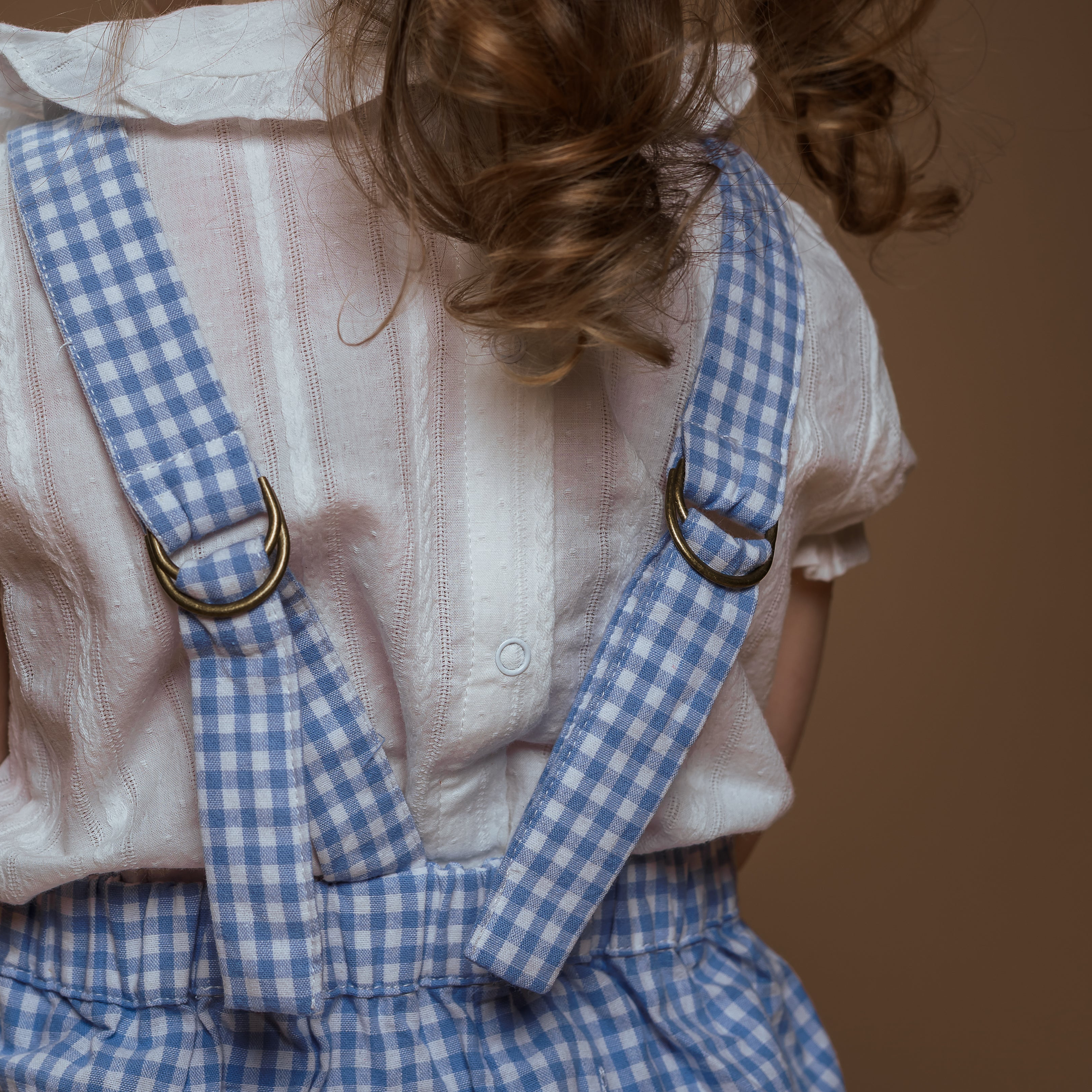 Child wearing a white shirt and blue checkered overalls on a brown background