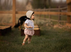 Baby in a bonnet and bloomers in a blurred field with dog in background