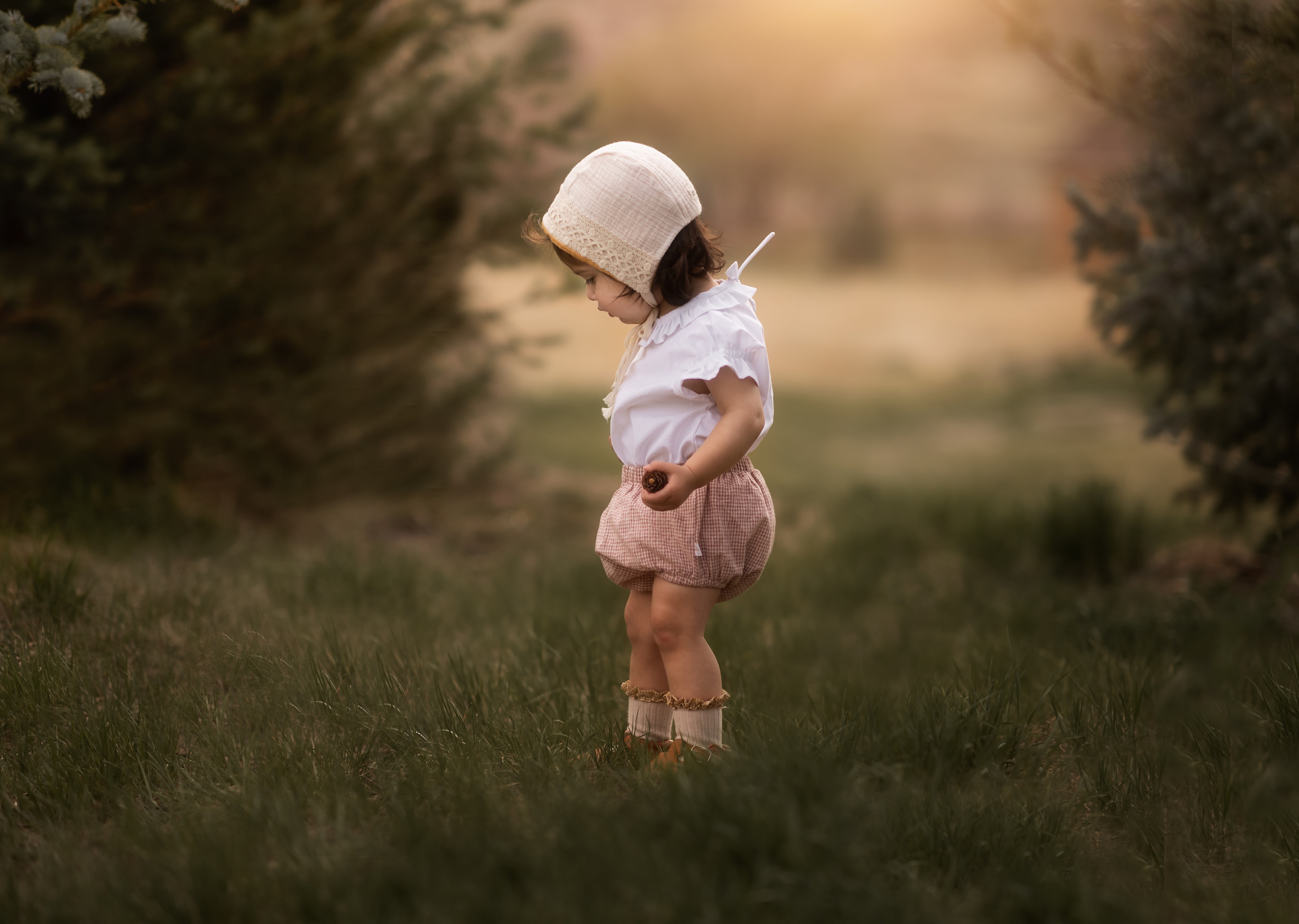 Sustainable rust gingham linen bloomers for babies, displayed in profile on a model girl wearing a sweet bonnet in a field of grass. Made from a soft cotton-linen blend, these breathable and lightweight bloomers are a wardrobe essential and pair beautifully with our dresses for an eco-friendly look.