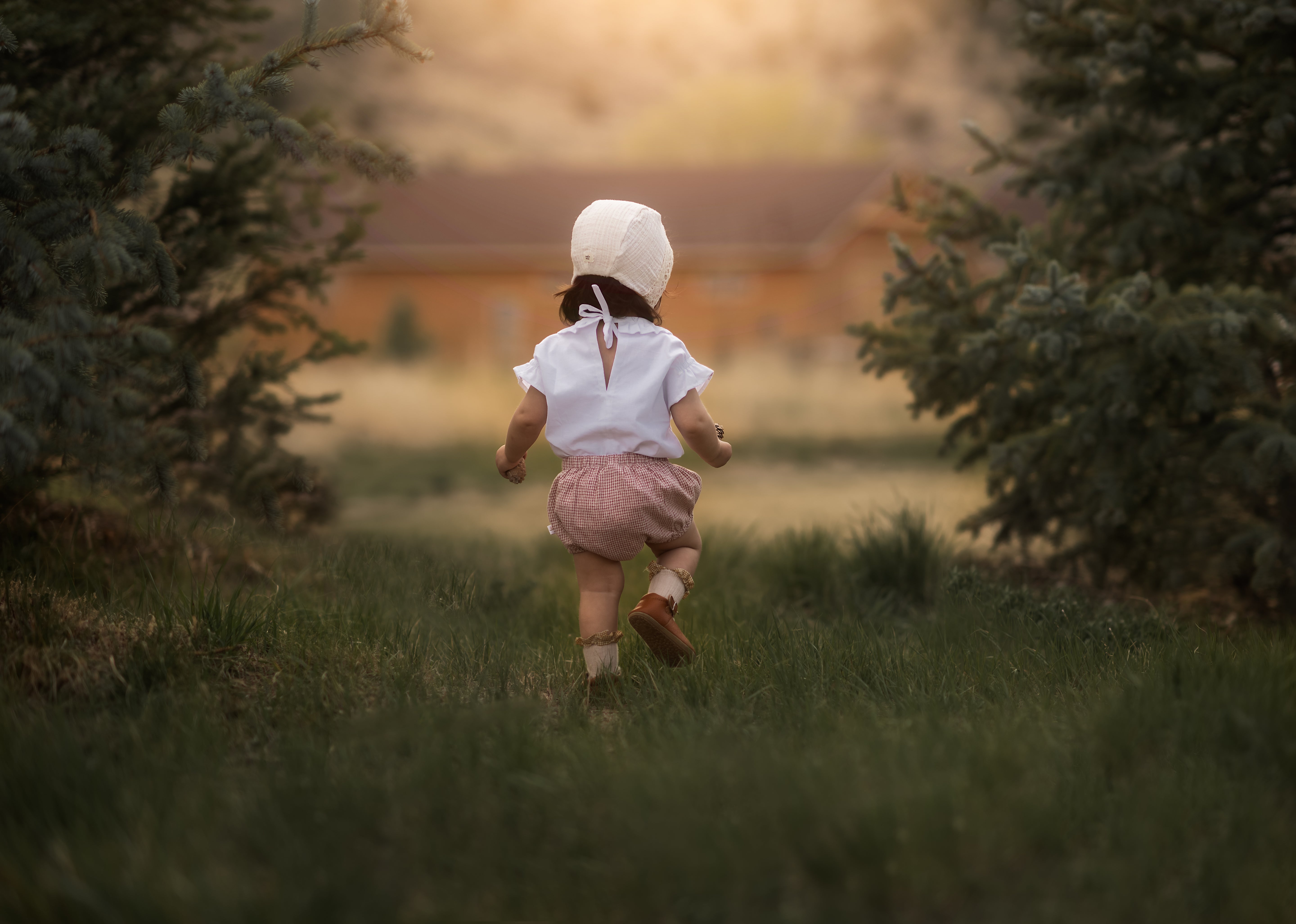 Charming rust gingham linen bloomers for babies, showcased from the back on a model girl wearing a delightful bonnet in a sunny field of grass. Made from a soft cotton-linen blend, these lightweight and breathable bloomers ensure all-day comfort and style, making them an essential piece for eco-friendly baby wardrobes.
