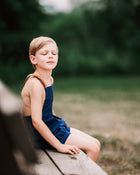 Boy with eye closed sitting on a bench wearing navy overalls