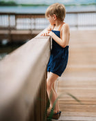 Boy in navy linen overalls by water