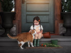 Child in checkered dress and green boots sitting on a step with an orange cat, flowers, and a door in the background.