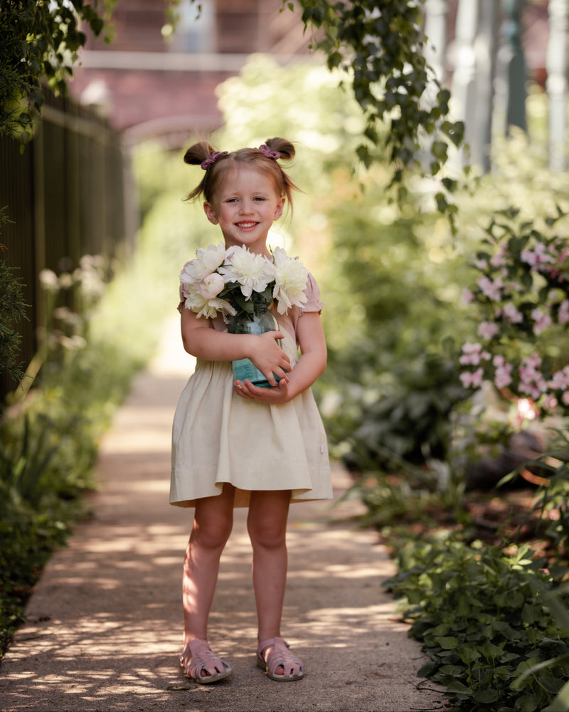 Young girl holding flowers in a garden setting wearing a natural linen pinafore