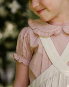 Close-up of a child wearing a beige blouse with ruffled collar and beige linen pinafore.