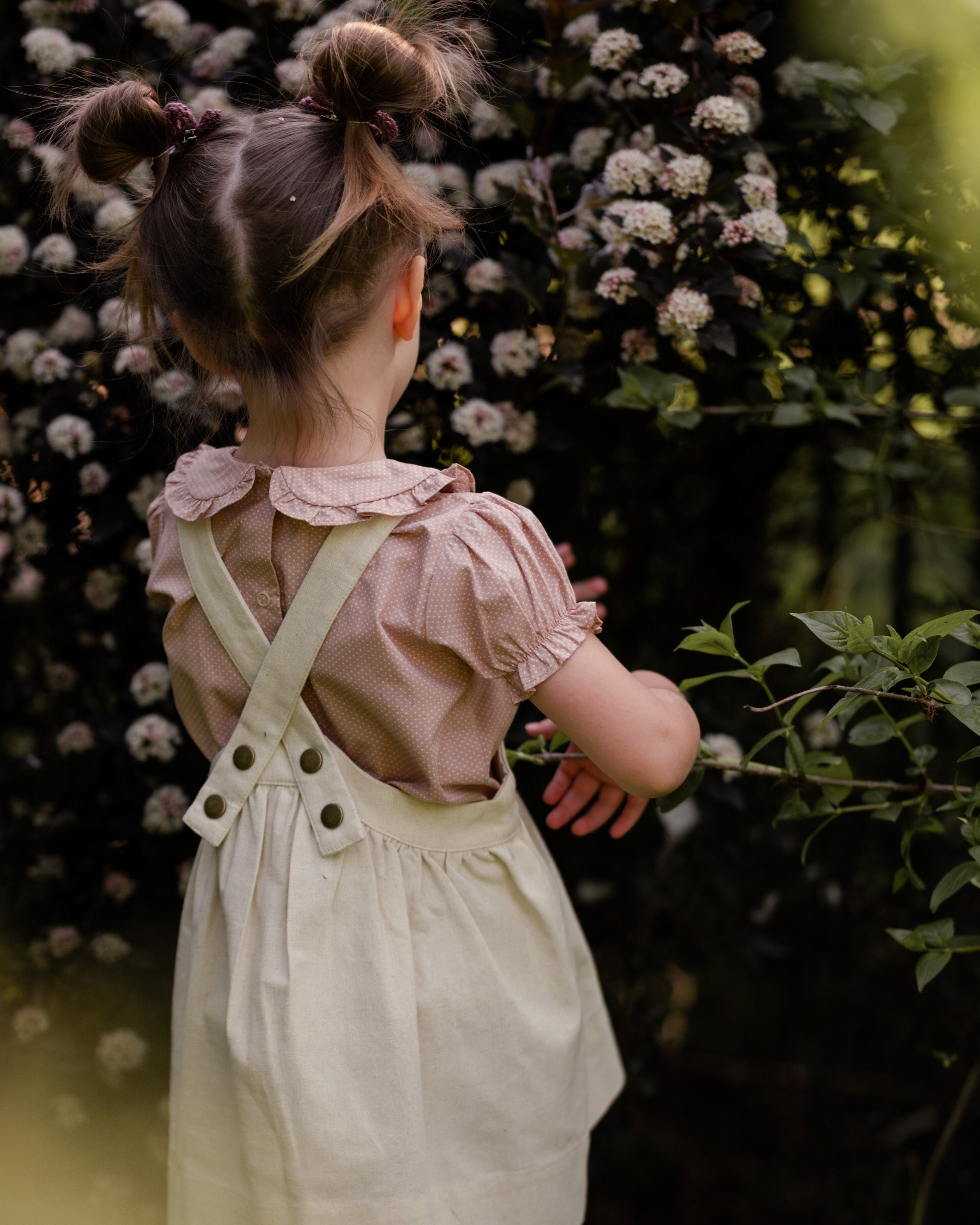 Girl wearing a beige blouse and beige pinafore with suspenders standing in front of a floral background