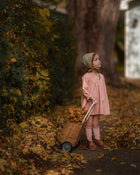 A young child wearing a pink dress, pink socks, and a patterned bonnet stands on a leaf-covered path next to a wicker cart filled with autumn leaves, with trees and bushes in the background.