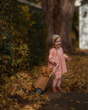 A young child wearing a dusty pink dress and a patterned bonnet pushes a small wicker cart filled with autumn leaves along a leaf-strewn path in a park.