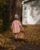 A young child seen from behind wearing a light pink coat and a patterned knit hat, pushing a wicker basket filled with dry leaves along a leaf-covered path in an outdoor autumn setting.