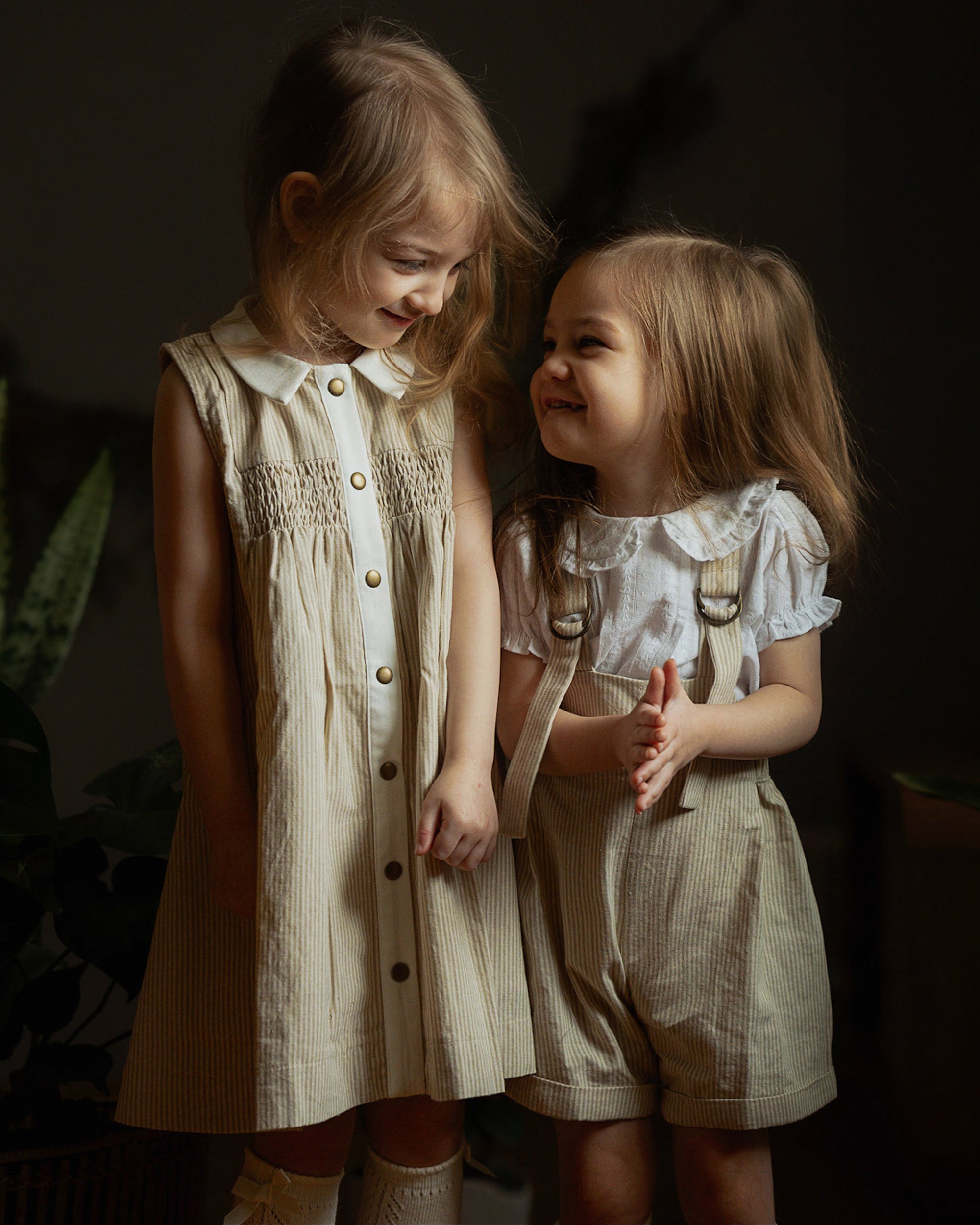 Two little girls wearing beige stripes Grow-With-Me Rompers and a matching beige stripes dress, playing together indoors, highlighting the adjustable straps, soft fabric, and coordinated timeless designs.