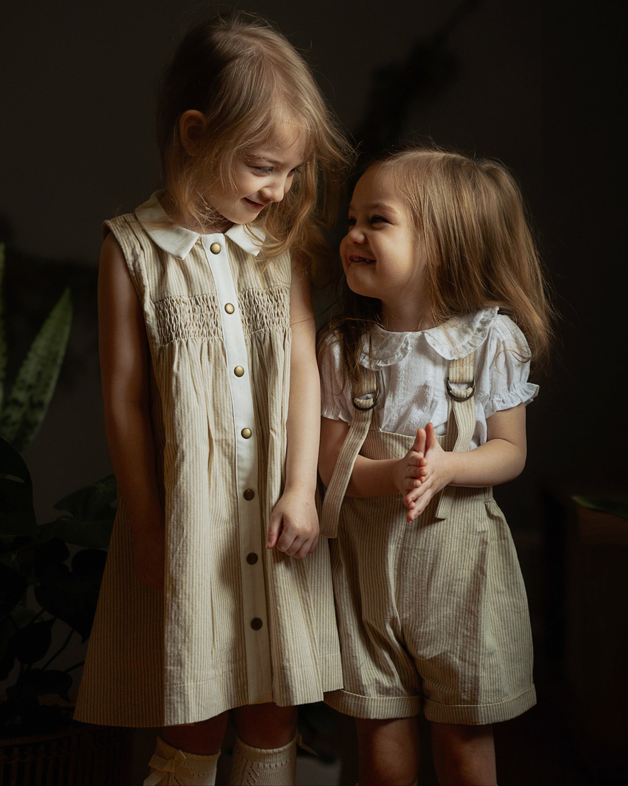 Two little girls wearing beige stripes Grow-With-Me Rompers and a matching beige stripes dress, playing together indoors, highlighting the adjustable straps, soft fabric, and coordinated timeless designs.