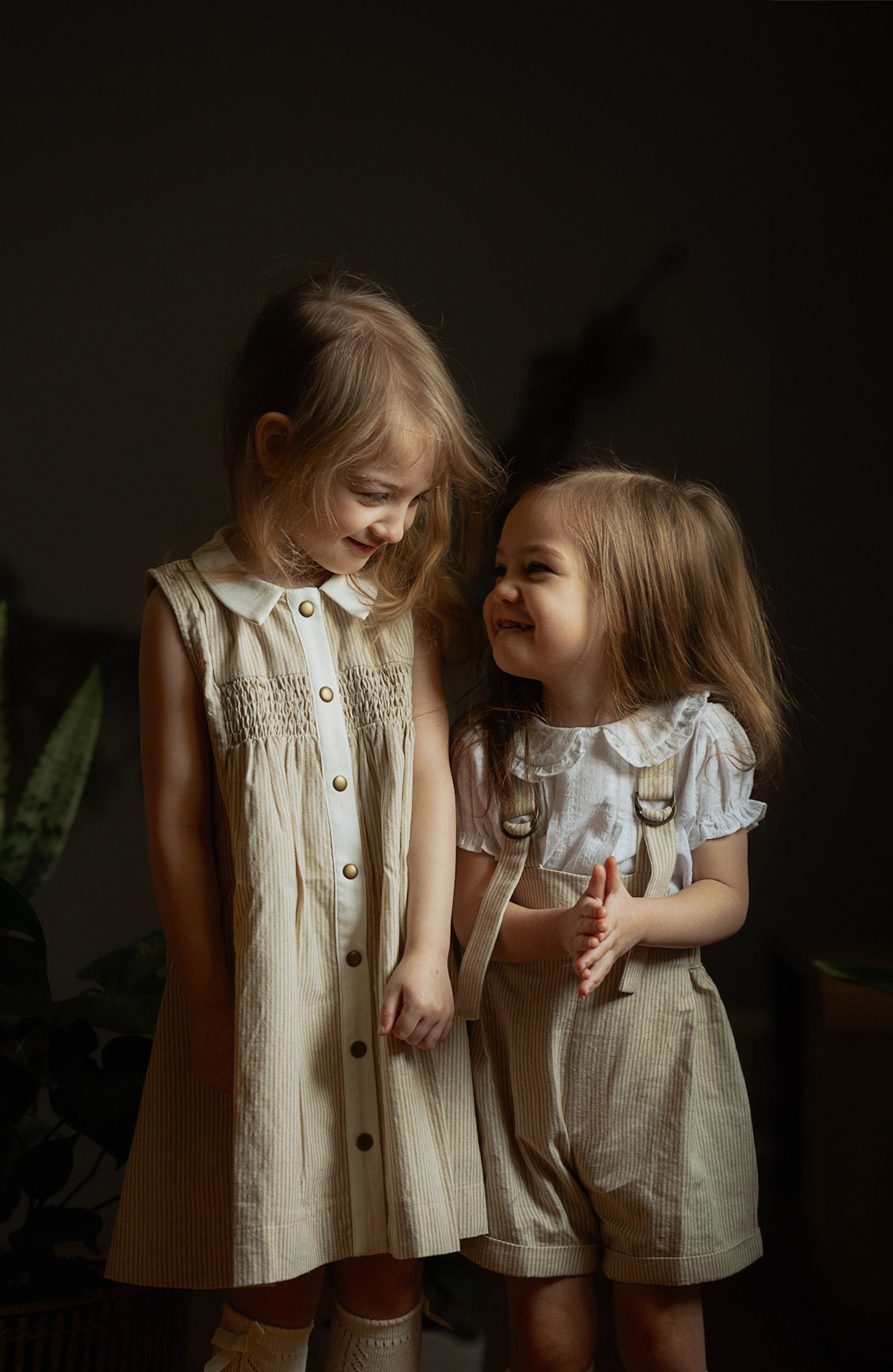 Two little girls wearing beige stripes Grow-With-Me Rompers and a matching beige stripes dress, playing together indoors, highlighting the adjustable straps, soft fabric, and coordinated timeless designs.