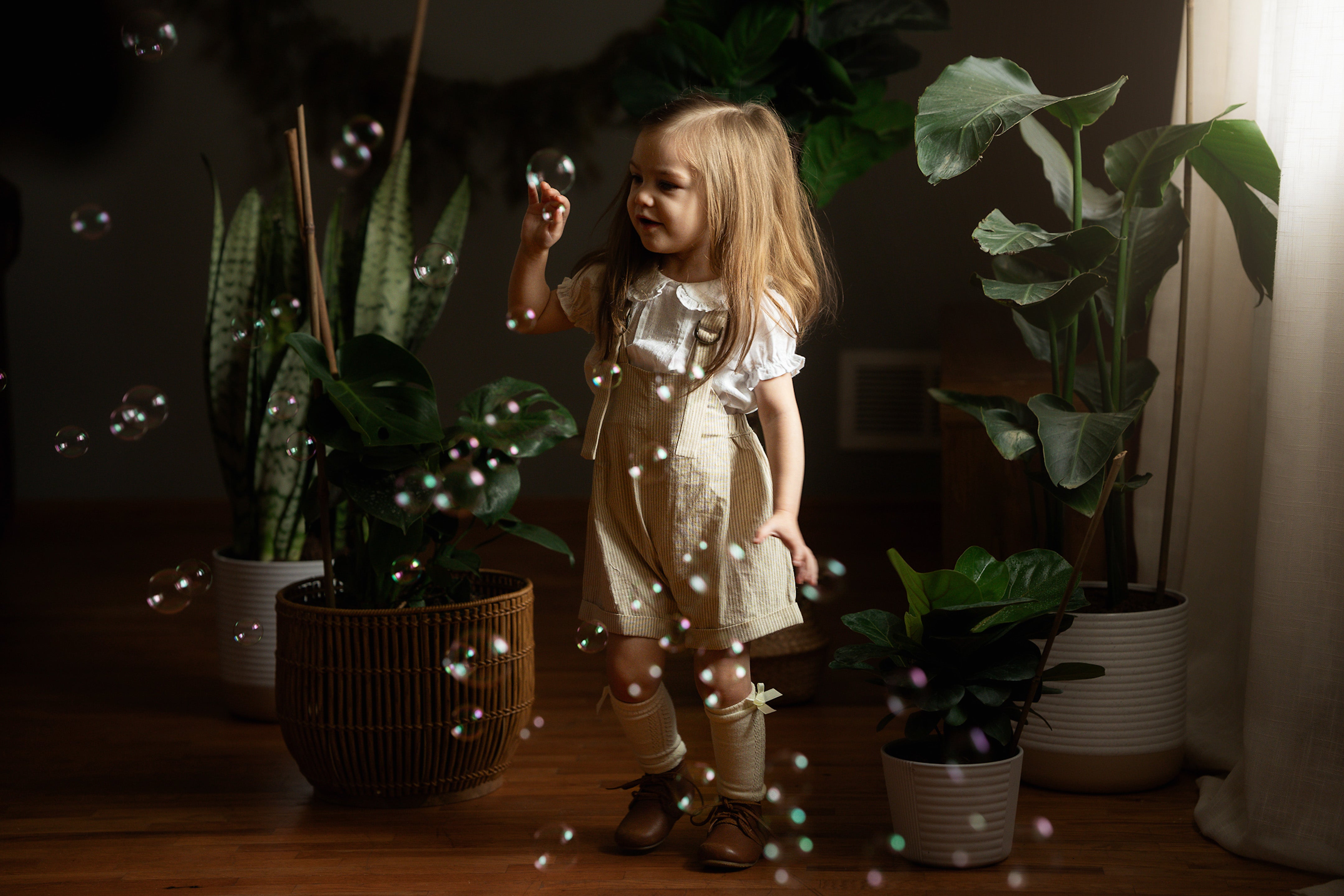 A tiny 2-year-old wearing a beige stripes Grow-With-Me Romper over a frilly white heirloom style blouse, playing indoors with soap bubbles in a cozy setting, showcasing the adjustable straps and soft fabric of the romper paired with the elegant details of the blouse.