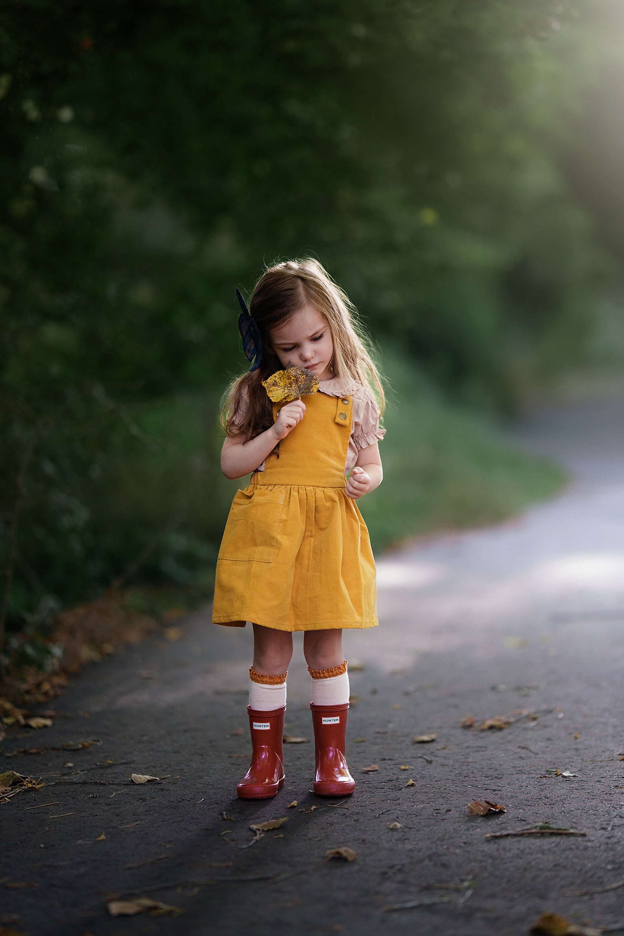 Model girl standing wearing rain boots and 3-year-old girl modeling the Mustard Pinafore paired with rain boots and a frilly beige dots blouse, highlighting a playful summer look that combines style and comfort.