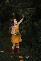 Side view of a girl in the Mustard Pinafore, joyfully picking flowers, demonstrating the garment’s versatility and how it can be worn on its own during summer adventures.