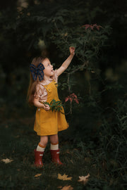 Side view of a girl in the Mustard Pinafore, joyfully picking flowers, demonstrating the garment’s versatility and how it can be worn on its own during summer adventures.