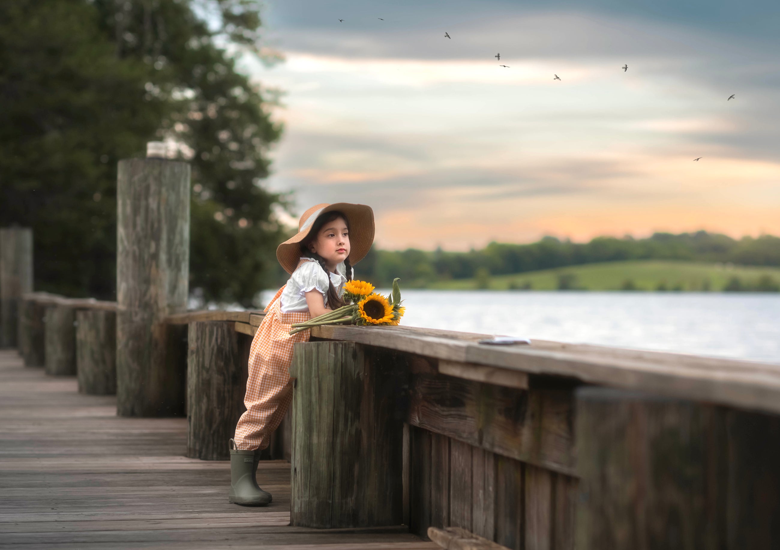 Girl on a wooden dock by a lake with sunflowers, wearing a hat and rain boots.